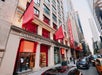 Street view of the Museum of Broadway building in New York City, with large red signs and banners, surrounded by other tall buildings and parked cars.