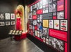 Exhibit with a red feathered gown on a mannequin, framed photos, documents in a display case, and a wall filled with historical posters, images, and text panels.
