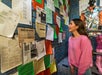 A woman in a pink sweater looks at a wall covered with various papers, posters, and documents in a brightly lit room with blue brick walls.