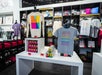 Display of colorful graphic t-shirts, mugs, and merchandise in a modern museum gift shop, with shelves of folded shirts and various souvenirs in the background.