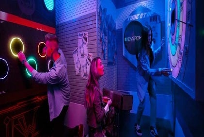 Three people interact with illuminated gaming stations in a dimly lit, futuristic room.