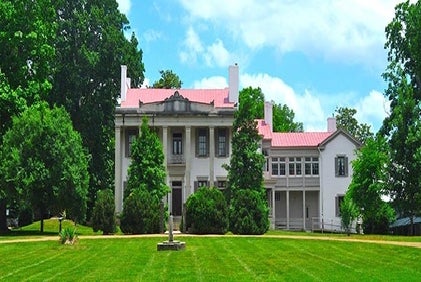 A large white historic mansion with columns, surrounded by green trees, sits under a partly cloudy sky.