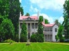 A large white historic mansion with columns, surrounded by green trees, sits under a partly cloudy sky.