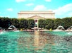 A large building with classical architecture is seen across a pond with fountains spraying water, surrounded by greenery and people.
