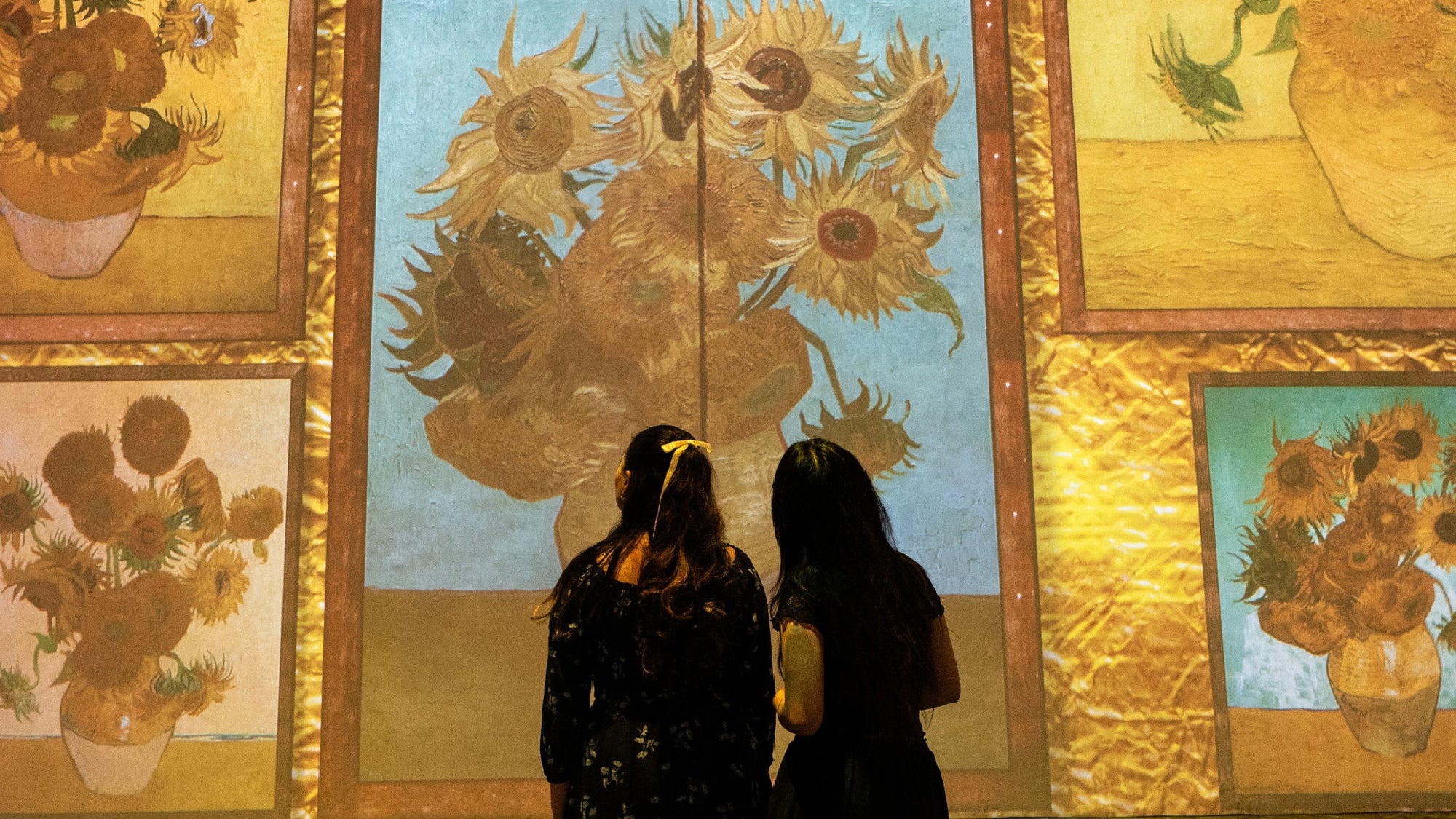 Two people stand facing large illuminated projections of Vincent van Gogh's sunflower paintings displayed on a wall.