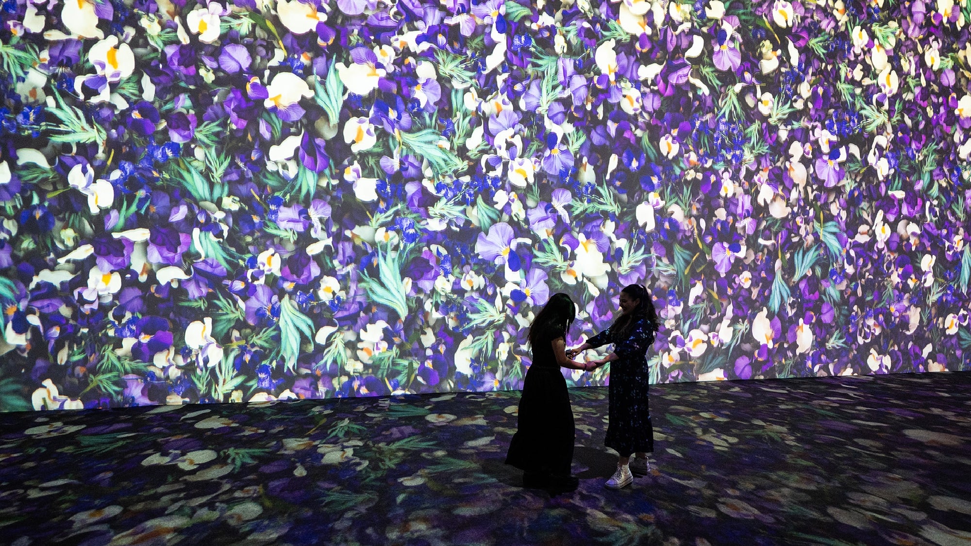 Two people stand in front of a large wall projection featuring densely packed purple, white, and yellow flowers with green foliage.