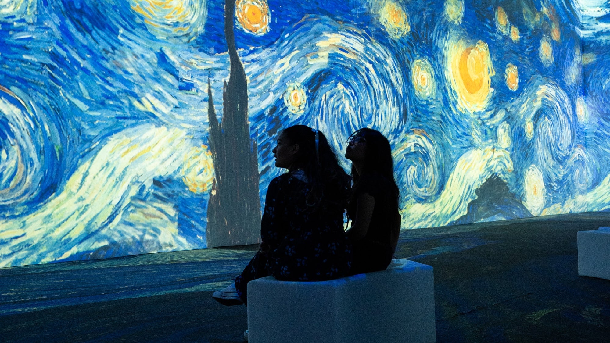 Two people sit on a bench, facing a large illuminated projection of van Gogh's "Starry Night" in a dark exhibition space.