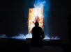 A person sits in front of a glowing, upright screen displaying a handprint amid bright, abstract light effects, surrounded by mist in a dark room.