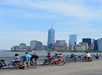 People stand and ride bikes along a waterfront promenade with the New York City skyline, including One World Trade Center, visible in the background on a clear day.