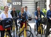 Six women wearing lanyards are standing with yellow bikes on a city sidewalk in front of a building marked "1370.