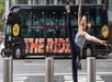 Person exuberantly posing with a high kick on a city street, with a large bus labeled "The Ride" and onlookers in the background.