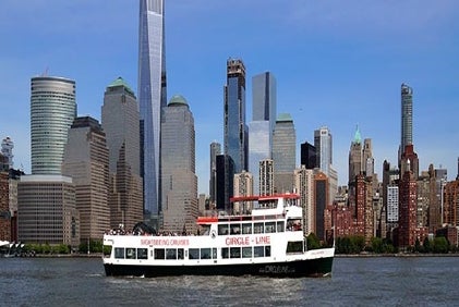 A sightseeing cruise ship sails on a river with a city skyline featuring modern skyscrapers and historic buildings in the background.