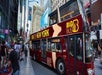 A maroon Big Bus New York tour bus pulling away from the curb with people walking on the side walk to the left.