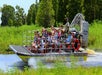 An airboat full of people splashing through the water with bright green weeds on their side of them on a sunny day.