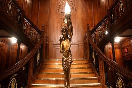 A gold statue of a cherub in the middle of a large wooden staircase at the Titanic - The Artifact Exhibition.