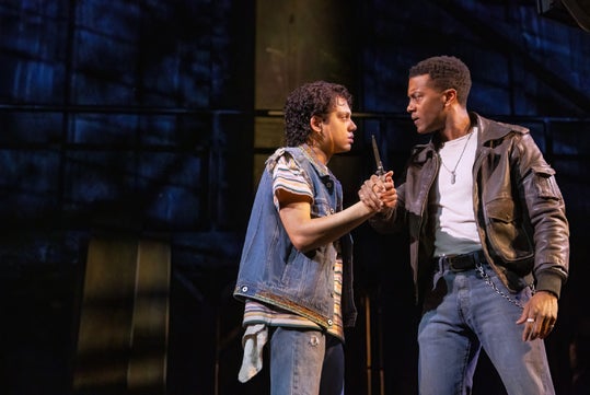 Two actors perform on stage, one handing a gun to the other. The scene is dramatic, with a dark background and intense expressions.