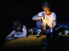 Two people are in a dimly lit setting; one person is lying on a blanket, looking at another person who is sitting, holding a lit flashlight over a book.