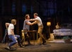 Three men perform an intense scene at a table, while another man sits in a car on a dimly lit stage set.