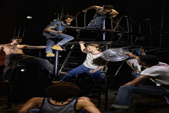 A group of men in casual clothing and jeans pose dynamically on a metal structure on stage, with one holding a round object.