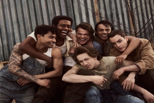 The cast of The Outsiders in costume huddled together laughing with a tin wall behind them.