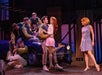 Stage scene with a group of young people in 1950s-style attire. Some sit on a car, while a man and woman stand close, and another woman watches. They wear varsity jackets and vintage dresses.