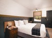 A hotel room with rustic decor, a king bed with a wooden headboard, a night stand with snacks and wine on it and window on the far side of the room.
