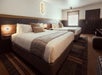 A hotel room with two queen beds with wooden headboards, ottoman, nightstand, desk with a TV on it and a window on the far wall.