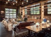 A lobby lounge area with rustic decor and plenty of seating around an indoor fire pit with large paneled windows all around the room.