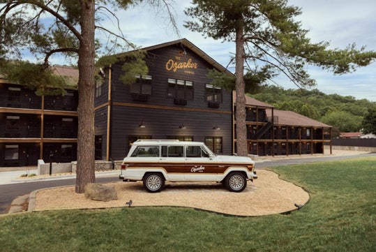 A vintage SUV is parked in front of a three-story building labeled "The Ozarker Lodge," surrounded by trees and grass.