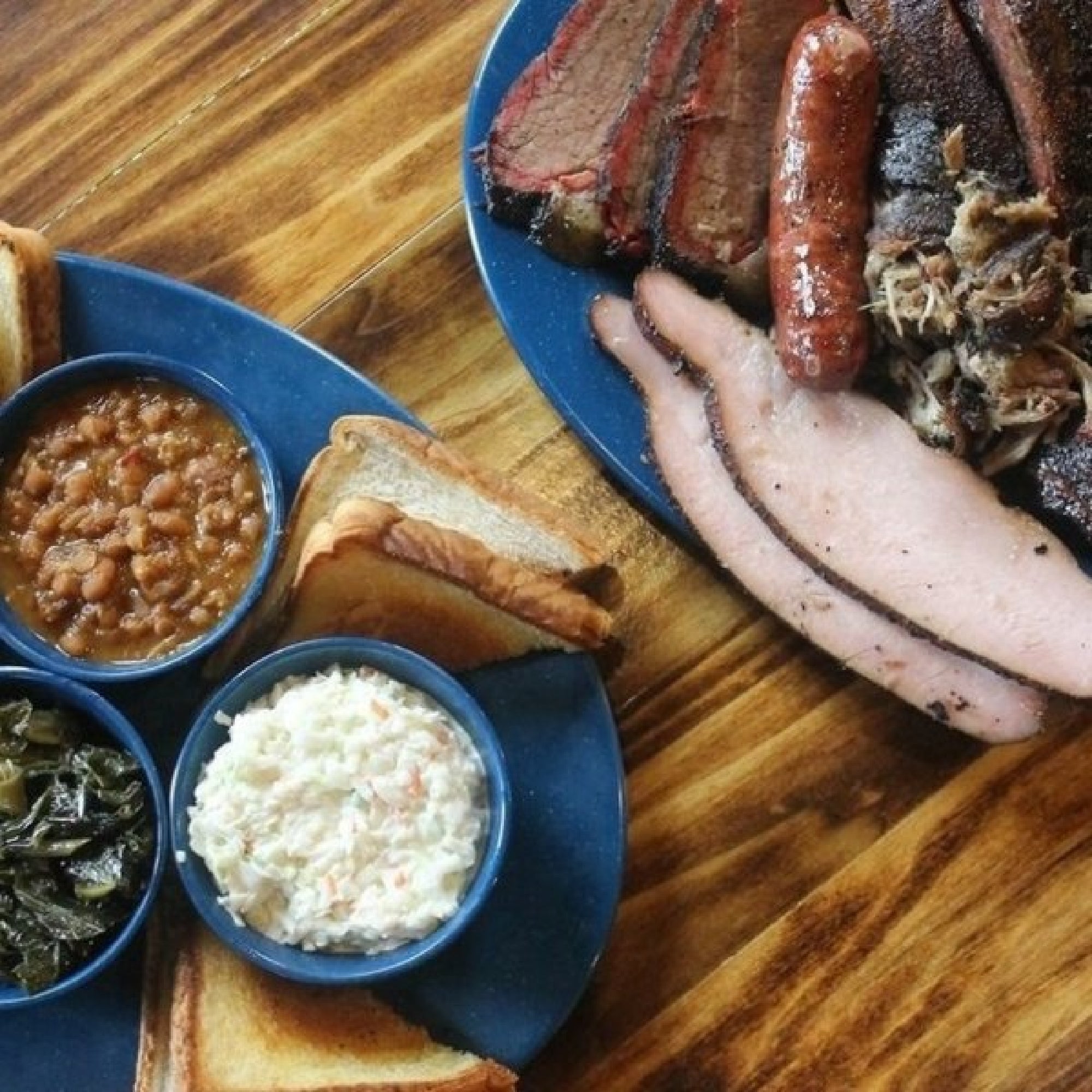 Plates of barbecue with sliced brisket, sausage, pulled pork, ham, baked beans, coleslaw, collard greens, and slices of toasted bread on a wooden table.