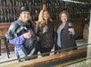 Three women stand indoors holding glasses of red wine, smiling at the camera. The background features a wall lined with wine bottles.