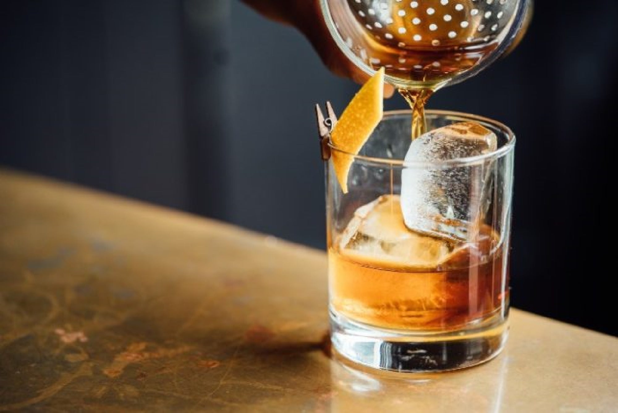 A hand pours a cocktail from a strainer into a glass with ice and a twist of lemon peel on a bar counter.