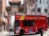 A red City Sightseeing Philadelphia bus drives past an ornate archway in an urban area. Passengers are visible on the upper deck, and buildings surround the street.