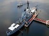 Aerial view of a large battleship docked at a pier with a yacht passing by in the background.