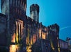 Historic stone castle with illuminated walls and a central round tower, set against a twilight sky.