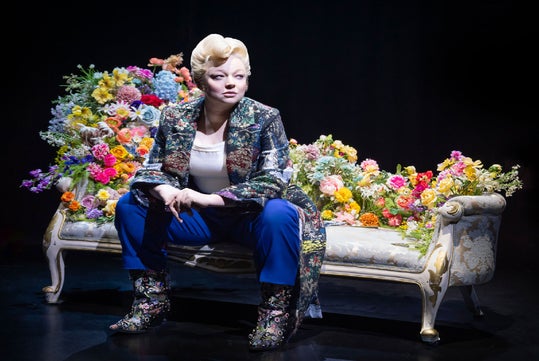 Person sitting on an ornate chaise lounge adorned with colorful flowers, wearing a patterned jacket, blue pants, and matching boots, against a dark background.