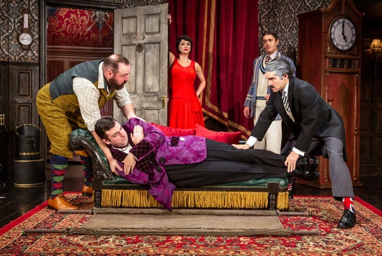A theatrical scene with actors in vintage costumes; one lies on a chaise lounge while others stand around, posing dramatically.