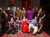 A group of performers in a theatrical pose on stage, with diverse expressions and clothing, set against an ornate background with rich colors.