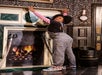 A person in overalls and patterned shoes reaches up near a fireplace, with ornate wallpaper and swords on either side.