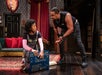 Two actors perform on stage. One is kneeling with a large box labeled "Mine! Keep off!!," while the other stands, gesturing animatedly. The setting resembles a living room with bookshelves.