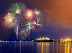 Colorful fireworks light up the night sky over a large cruise ship in a harbor, with city lights reflecting on the water.
