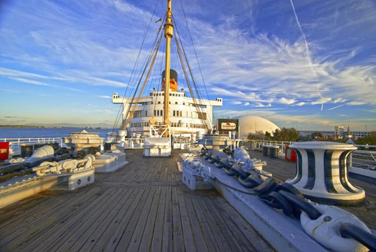 View from the deck of a docked ship with large anchor chains and a clear blue sky. A modern building and water are visible in the background.