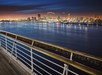 City skyline at night with illuminated buildings reflecting on the water, viewed from a pier with metal railings.