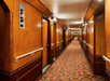 Long, carpeted hallway with wooden panel walls and multiple doors on each side, lit by overhead lights.