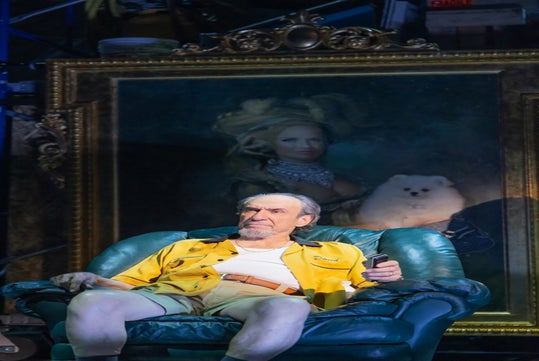 An older man in a yellow shirt sits in a green armchair holding a remote, with a large portrait of a woman and a white dog behind him.