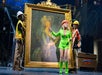 A woman in a neon green outfit and orange hard hat stands in front of a large framed portrait, flanked by two construction workers on a stage.