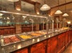 A buffet-style food counter features various hot dishes in metal trays, including sausages, eggs, and potatoes, with a glass sneeze guard and overhead lighting.