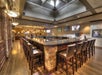 A spacious bar area with a central granite countertop, surrounded by cushioned wooden chairs. The room has wooden flooring, multiple screens, and modern pendant lights. Ceiling windows provide natural light.