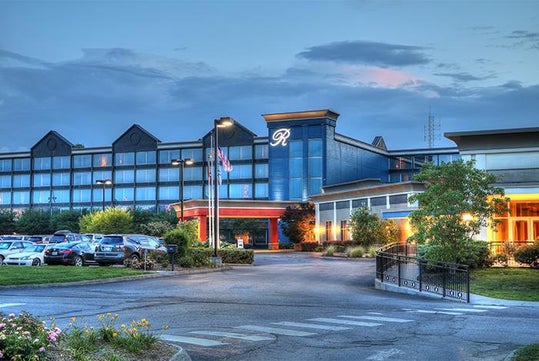 Exterior of a large hotel at dusk with a parking area in front, multiple windows, and a prominent letter "R" on the building.