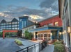 Hotel entrance with modern architecture, featuring large windows and mixed colors of blue, red, and beige. A covered driveway and lantern-style lights are visible. Cloudy sky in the background.
